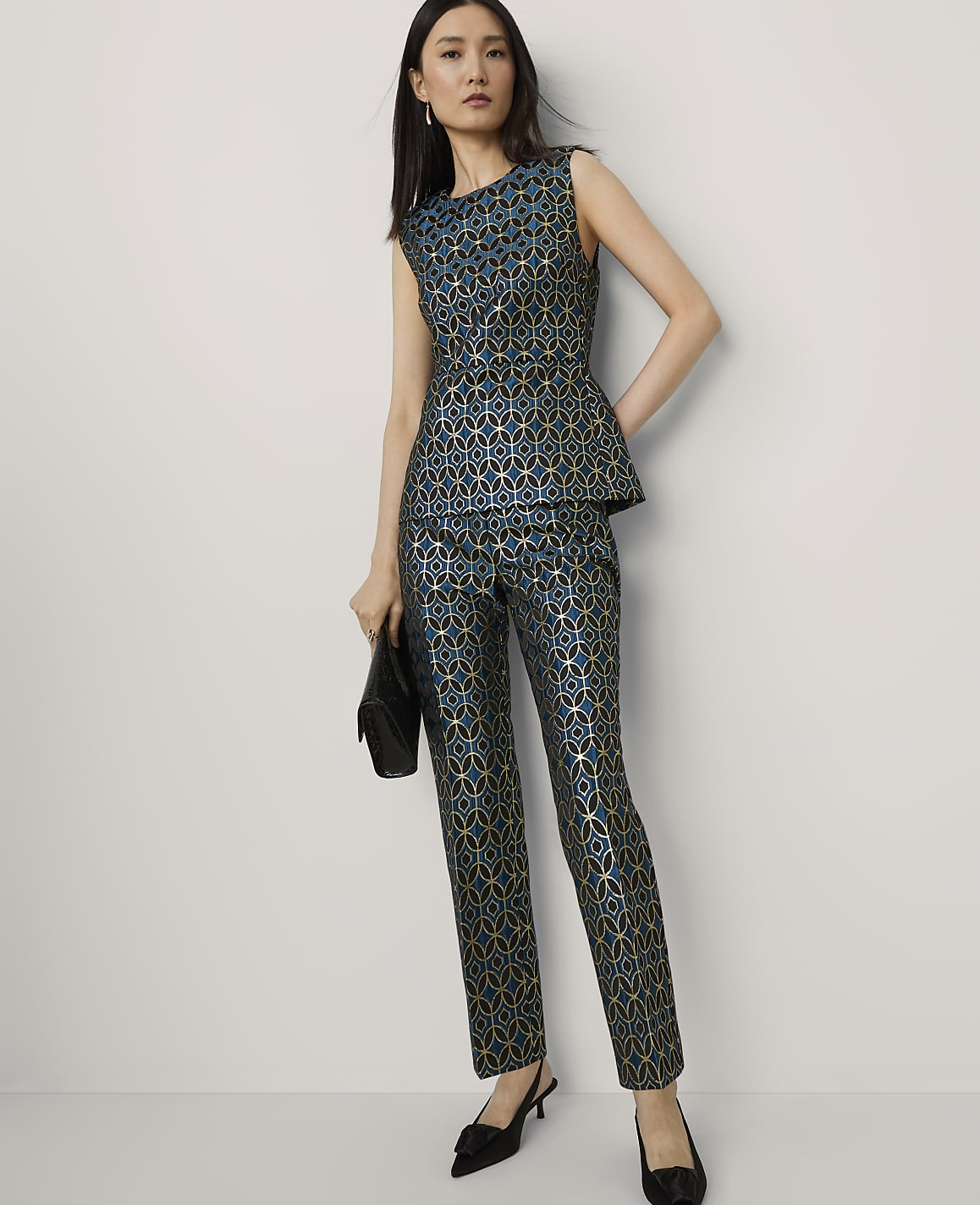 A woman in a teal and gold patterned peplum top and matching pants stands with a black clutch against a plain white background.