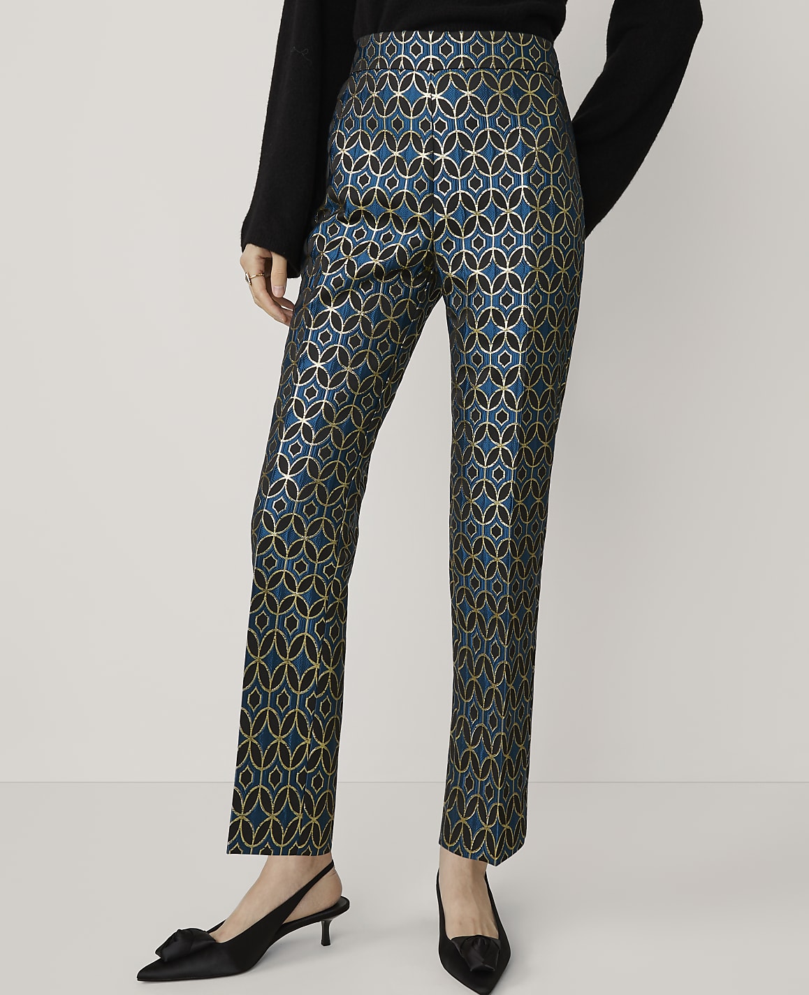 A person stands in the foreground wearing teal and gold patterned pants and black kitten heels, against a plain white background.