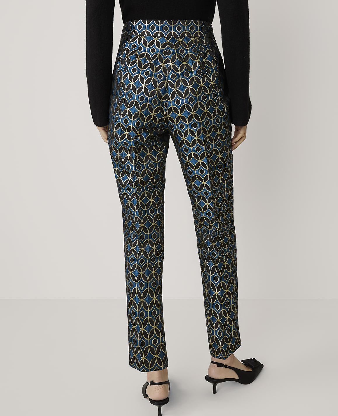 A person stands in front of a plain white background, wearing patterned teal and gold trousers and black heels.