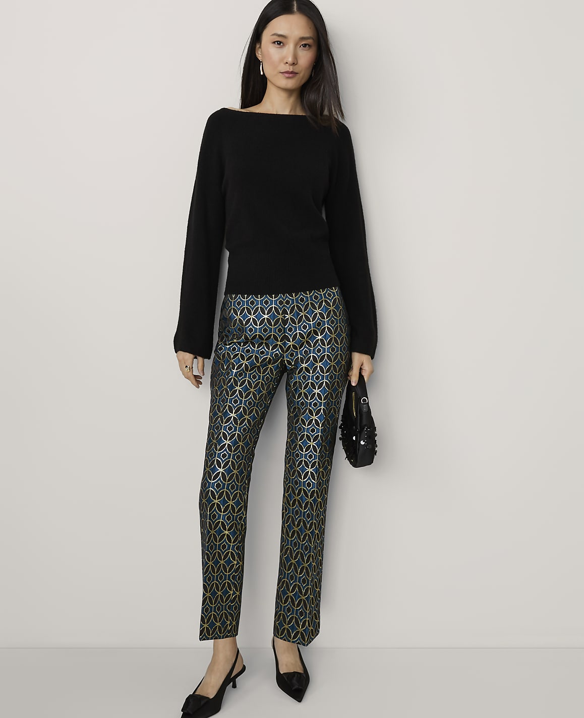 A woman in a black sweater and patterned pants stands against a plain background.