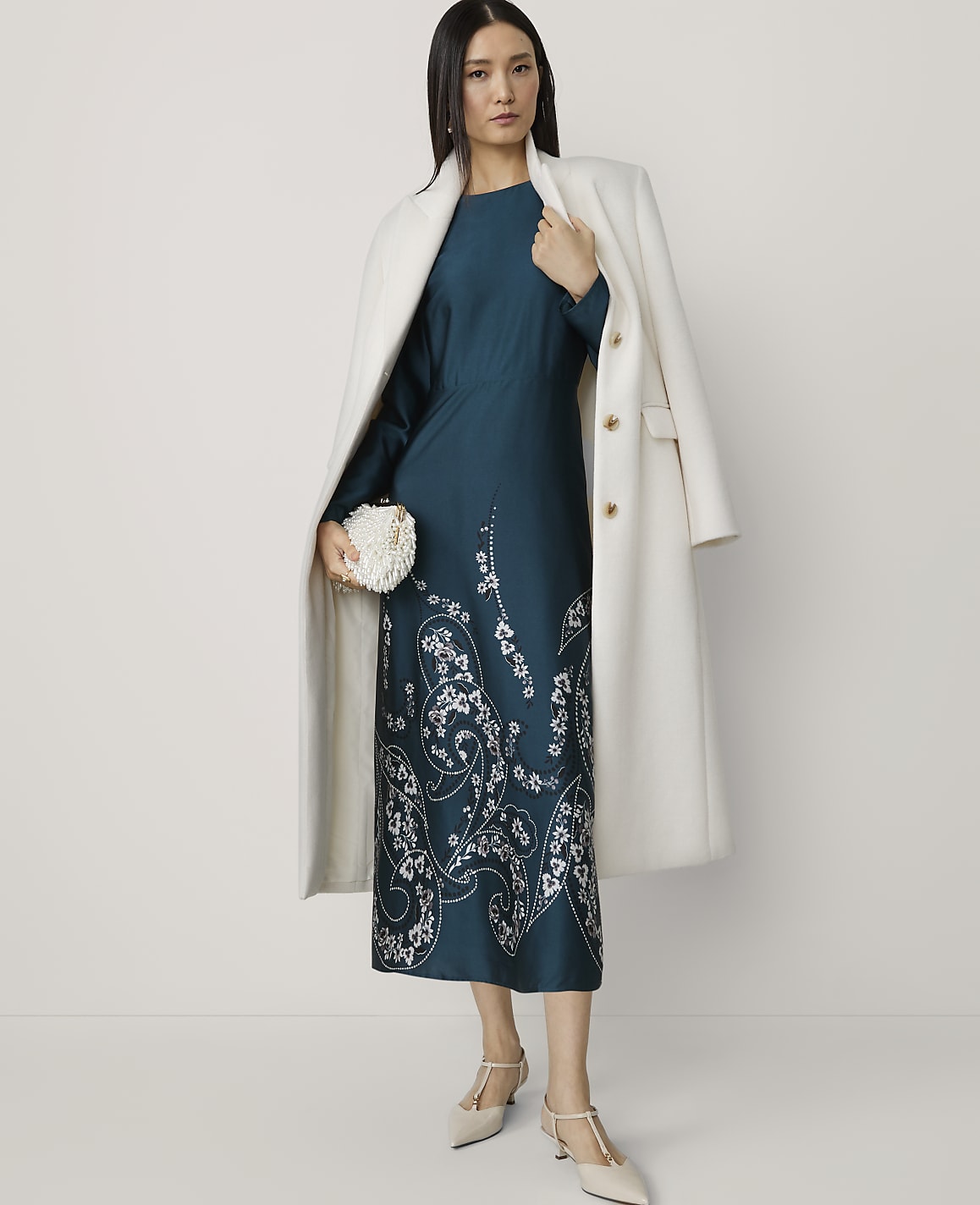 A woman in a teal dress with white floral embroidery and a white coat stands against a plain white background, holding a white clutch.