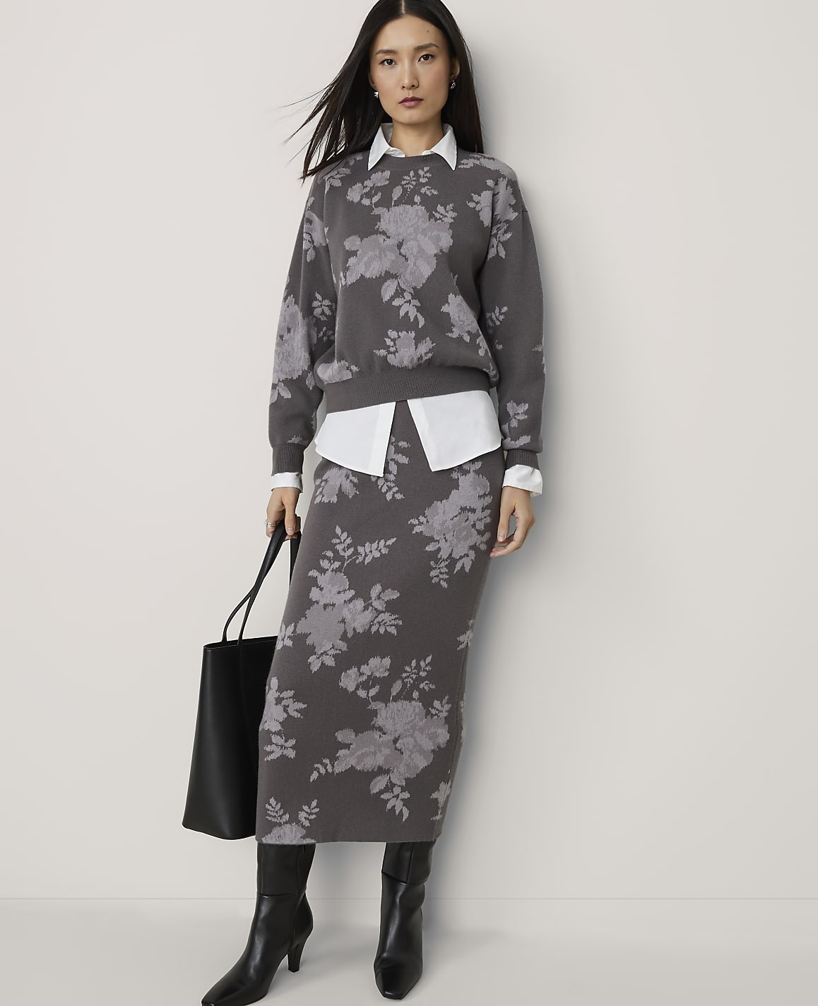 A woman in a floral sweater and skirt stands against a plain background, holding a black tote bag.