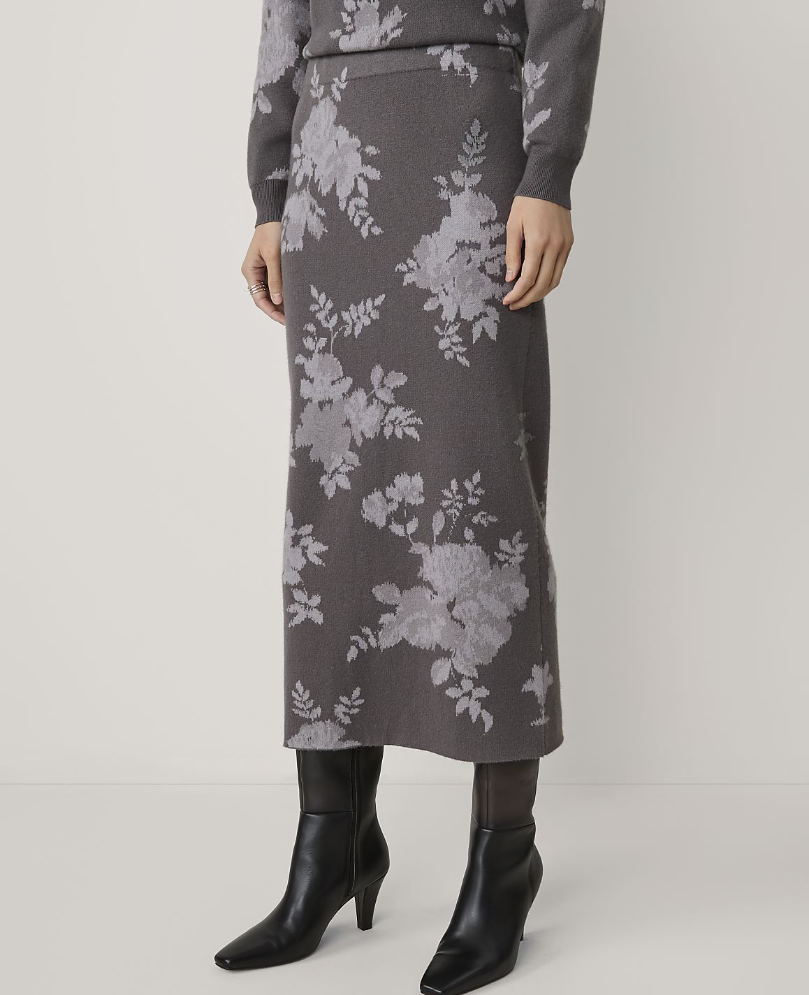 A person stands wearing a floral-patterned skirt and sweater, with black boots, against a plain white background.