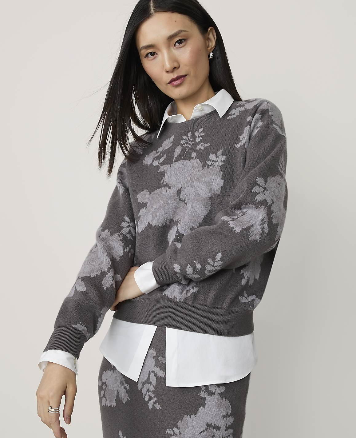 A woman in a floral sweater and white shirt stands against a plain gray background.