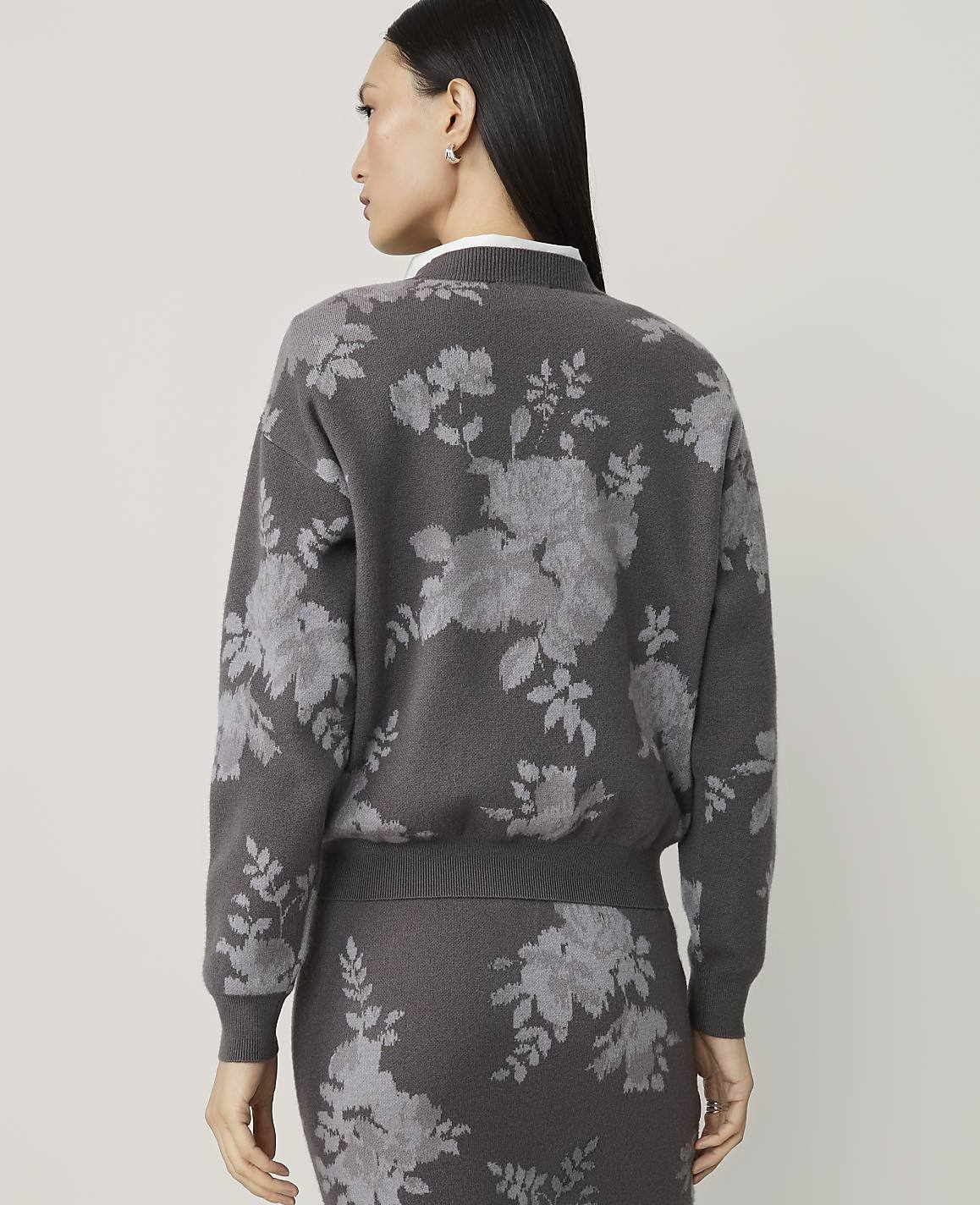 A woman in a gray floral-patterned sweater and skirt stands against a plain gray background.