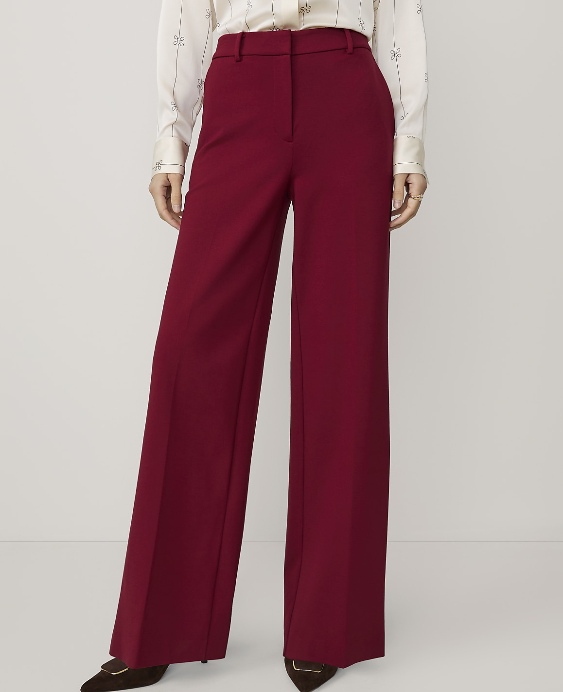 A person stands in front of a plain white background, wearing high-waisted, wide-leg burgundy pants and a patterned blouse.