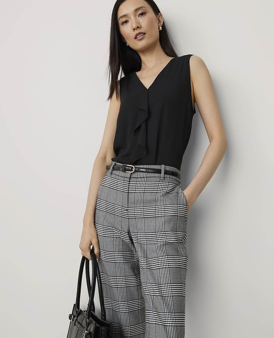 A woman in a black ruffled blouse and plaid pants stands against a plain white background, holding a black handbag.