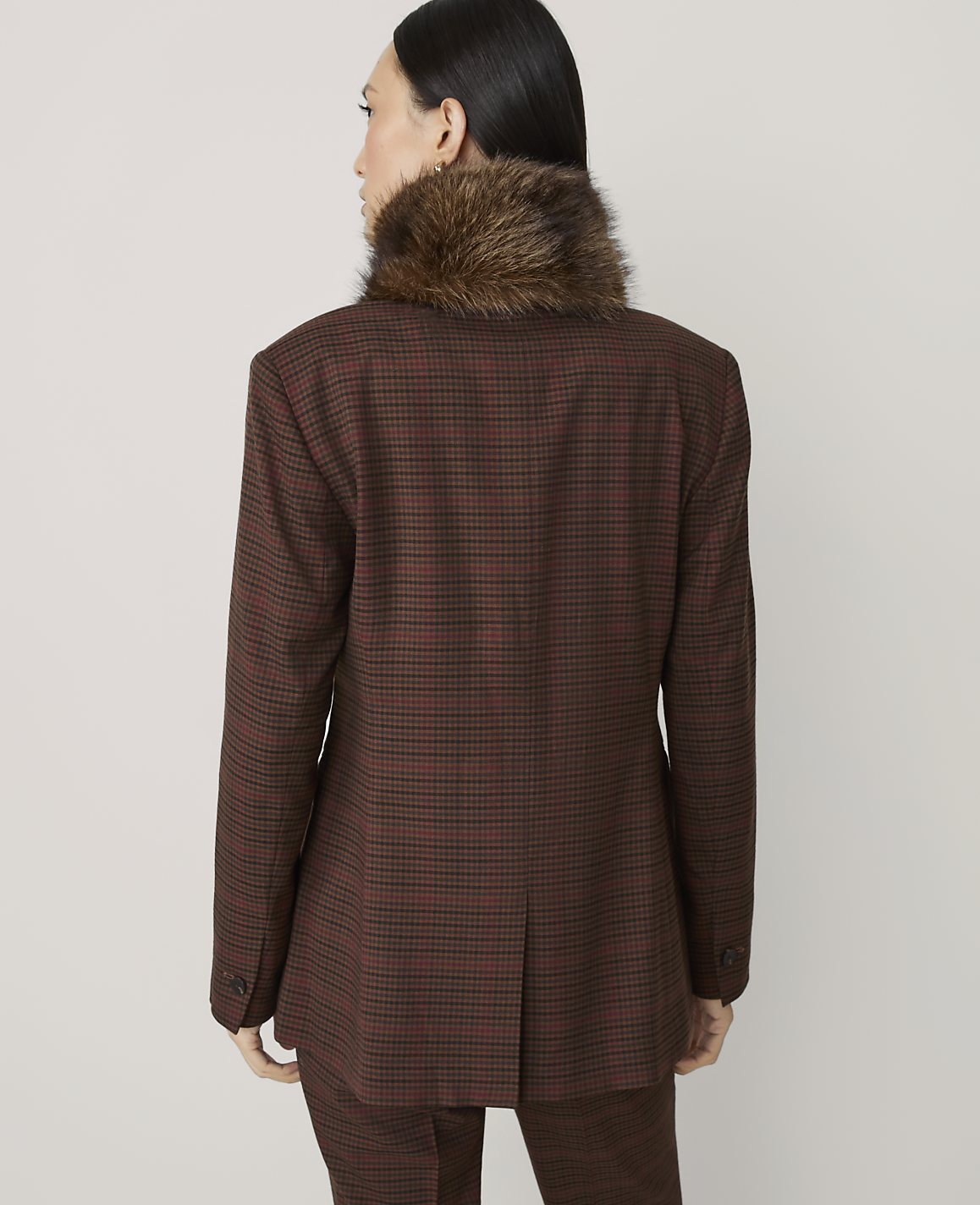 A woman in a brown plaid blazer with a fur collar stands against a plain, light gray background.