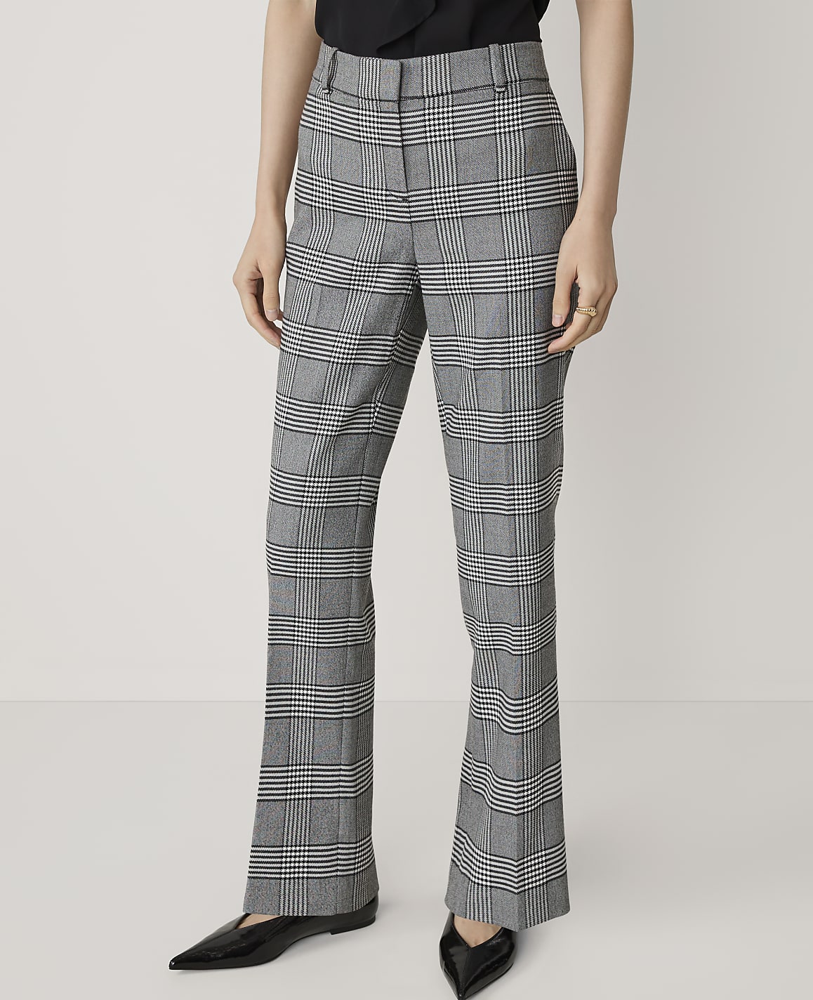 A person stands in front of a plain white background, wearing plaid flared trousers and black pointed-toe flats.