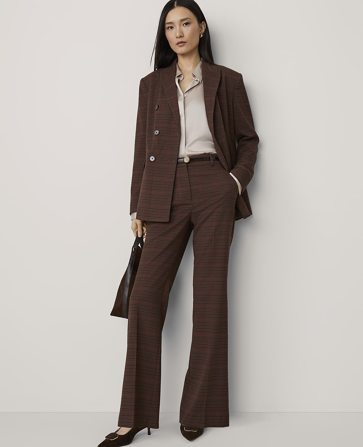 A woman in a brown plaid suit stands against a plain, light-colored background, holding a dark handbag.