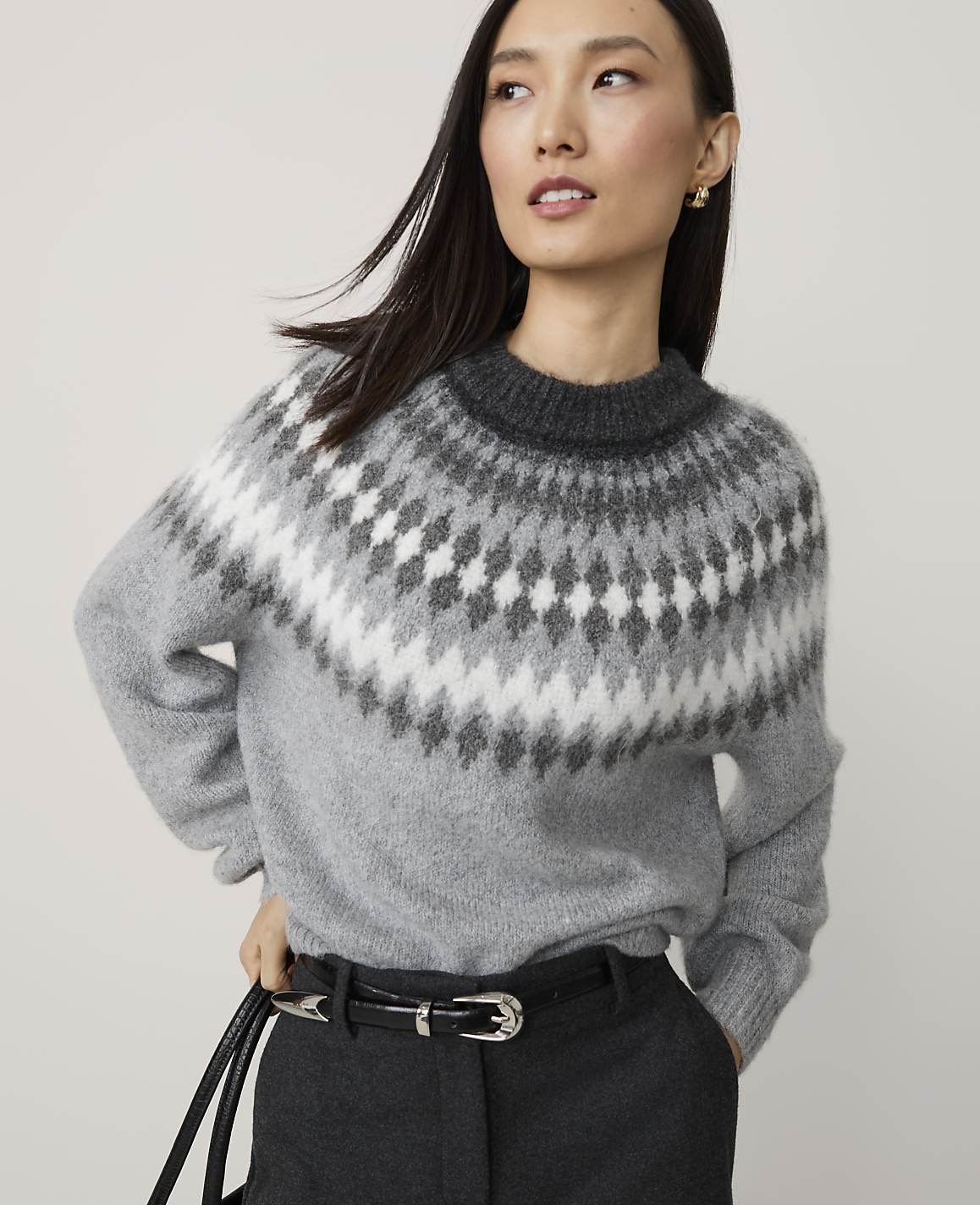 A woman in a grey fair isle sweater and black belt stands against a plain, light grey background.