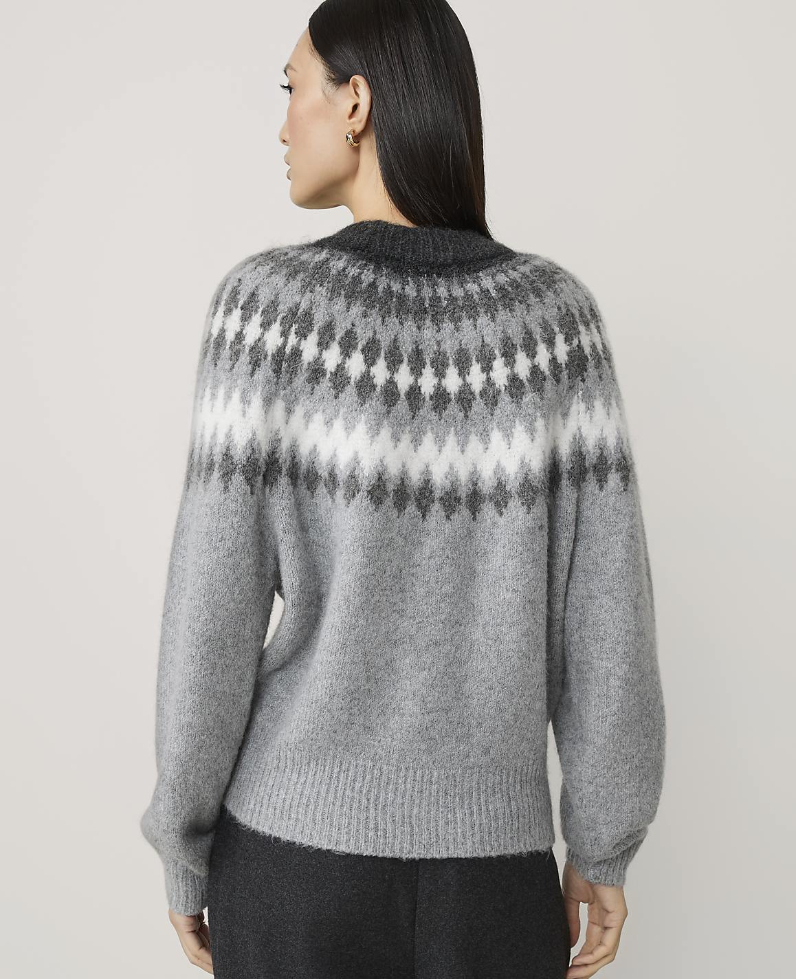A woman in a grey sweater with a fair isle pattern stands against a plain white background.