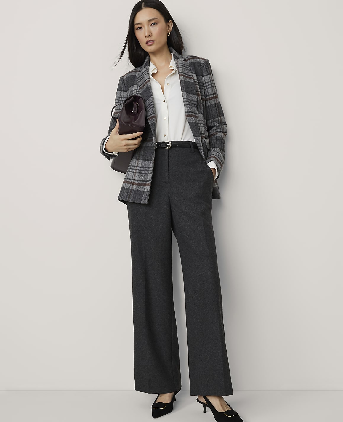 A woman in a plaid blazer and wide-leg pants stands against a plain white background, holding a burgundy handbag.