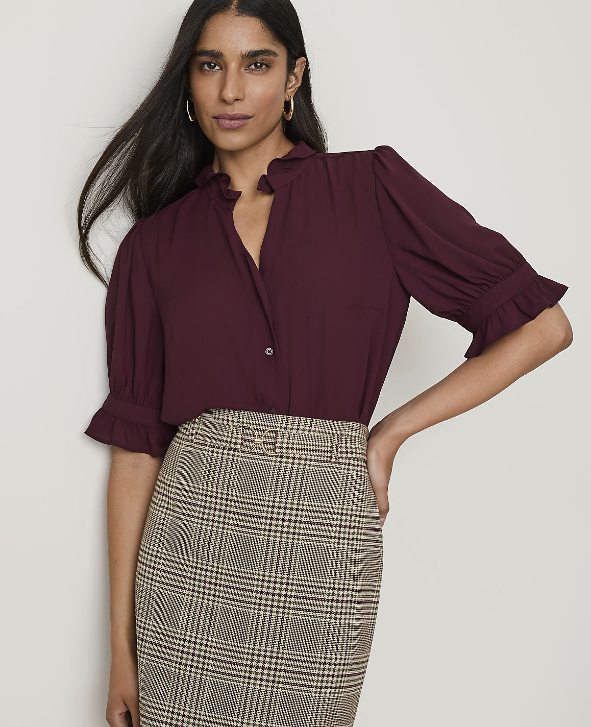 A woman in a burgundy ruffled blouse and plaid skirt stands against a plain white background.