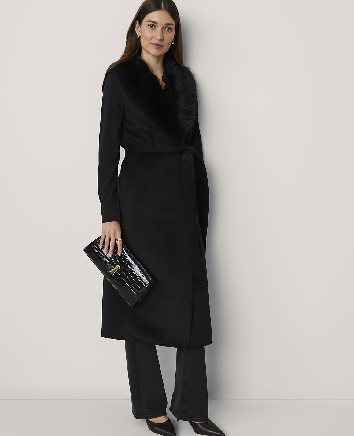 A woman in a black coat with a fur collar and a clutch purse stands against a plain white background.
