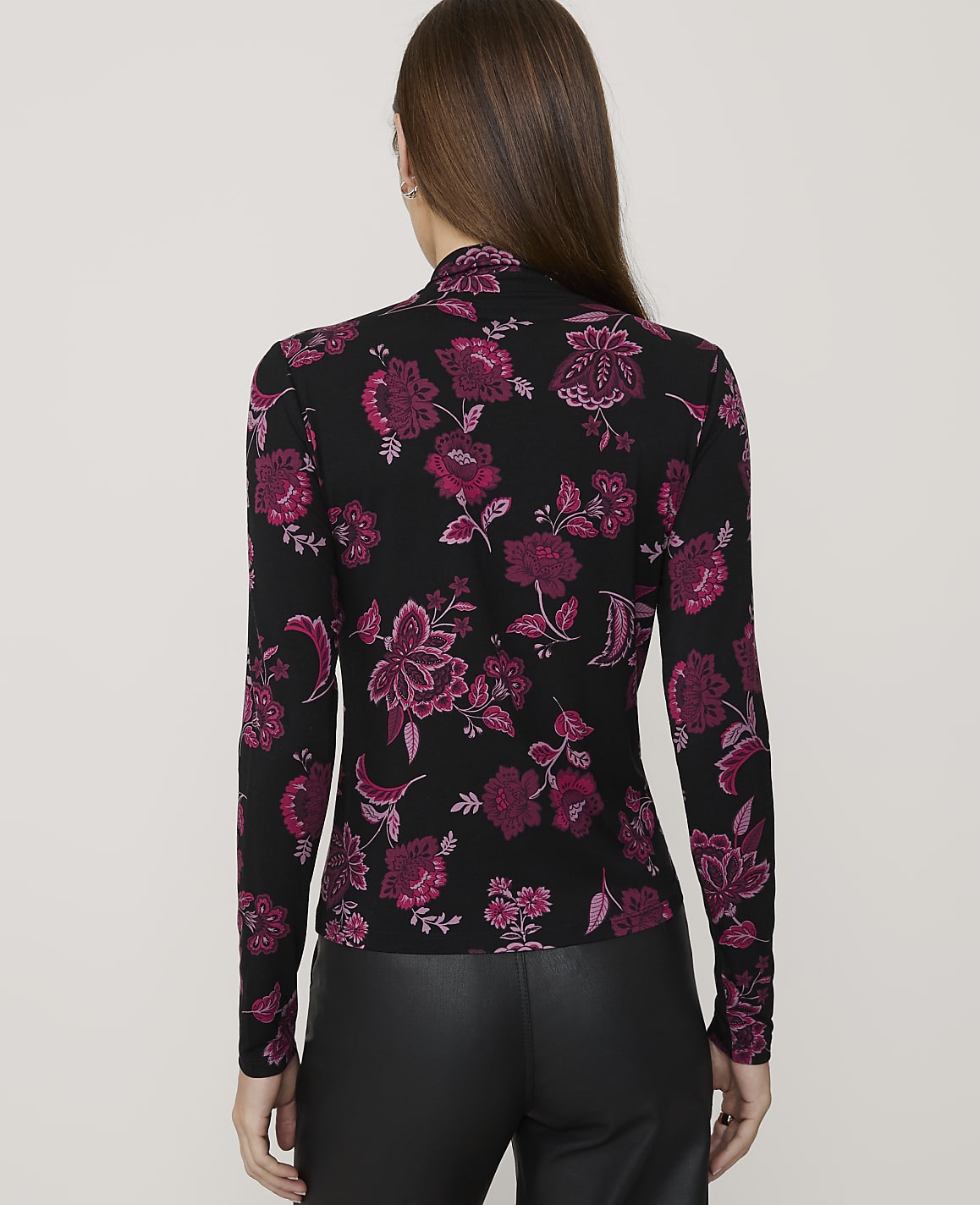 A woman in a black floral top and leather pants stands against a plain gray background.