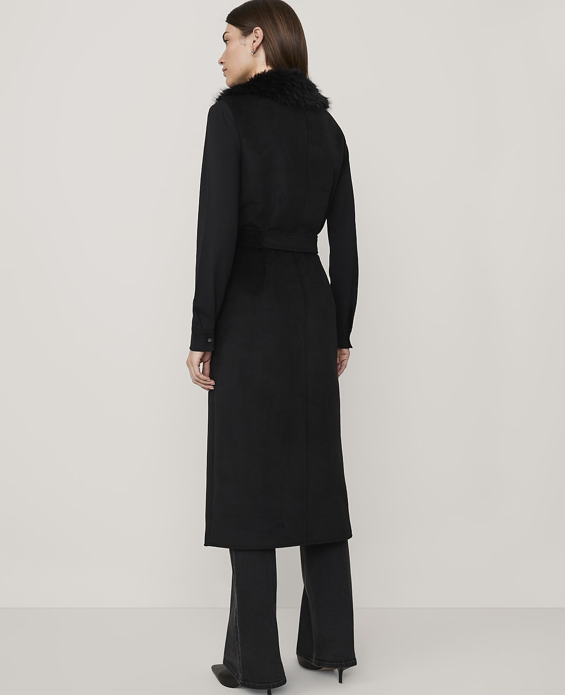 A woman in a long black coat with a fur collar stands against a plain, light gray background.