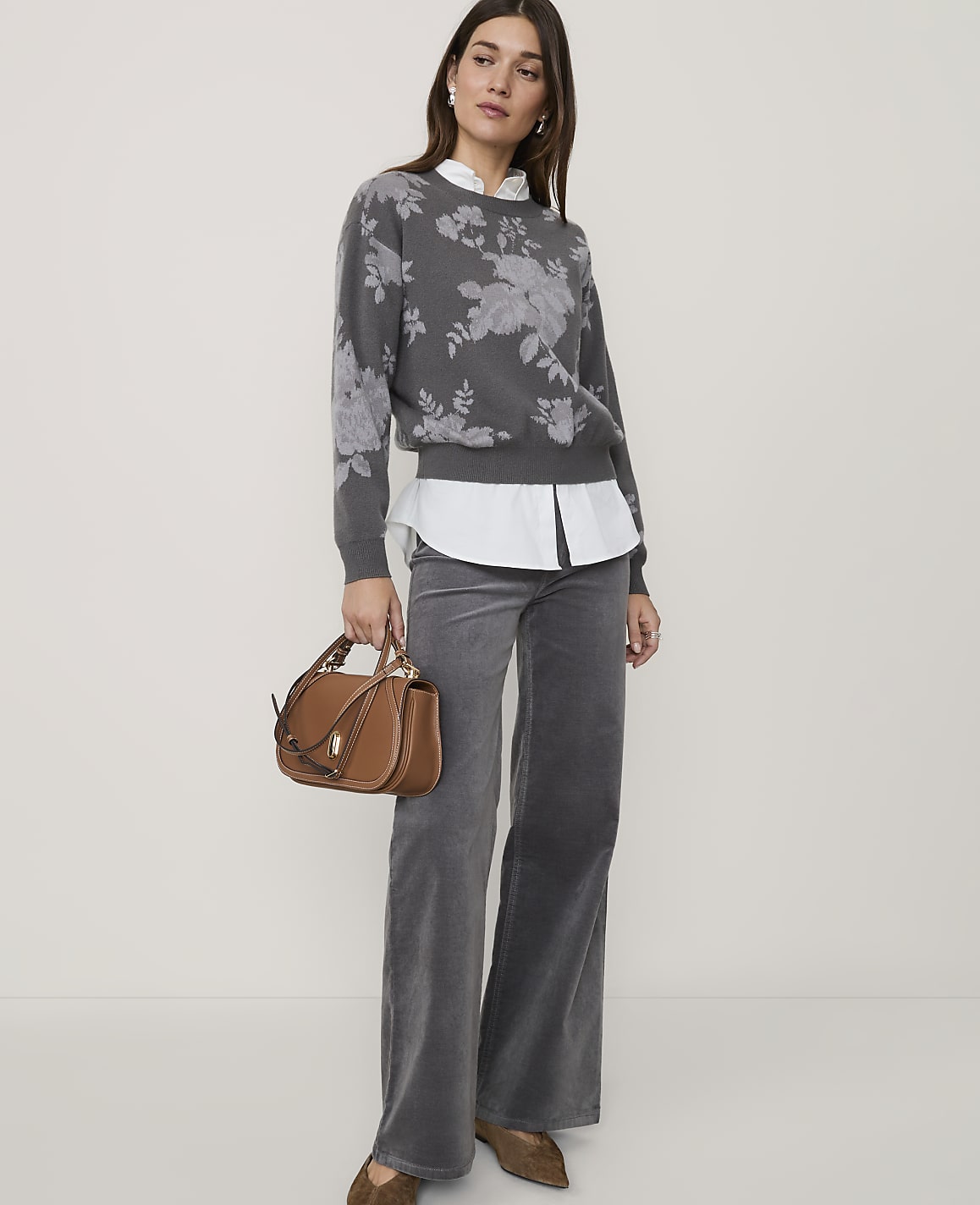 A woman in a floral sweater and gray pants stands with a brown handbag in front of a plain background.