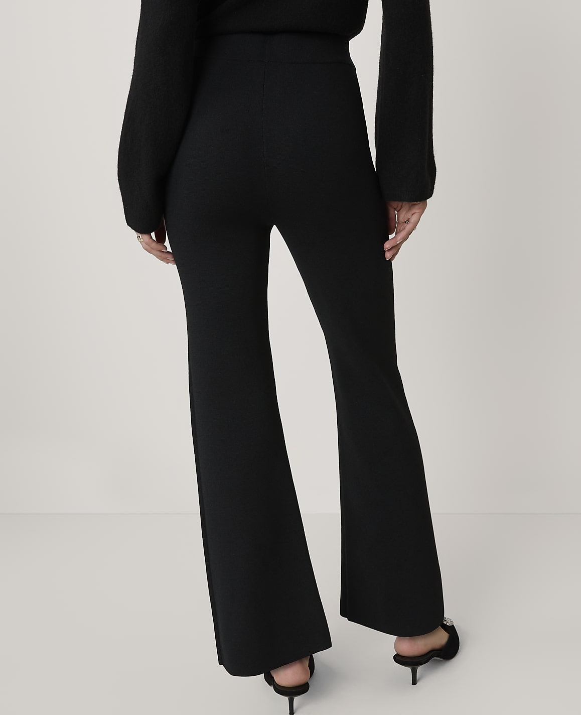 A person wearing black flared pants and a black sweater stands against a plain white background.