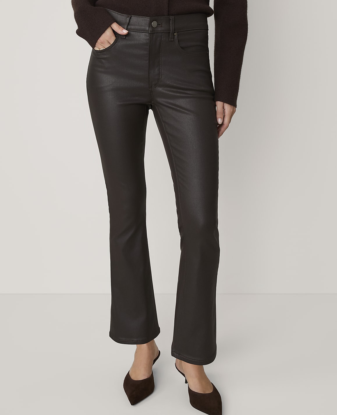 A person in dark brown, high-waisted, flared pants and brown mules stands against a plain, light brown background.