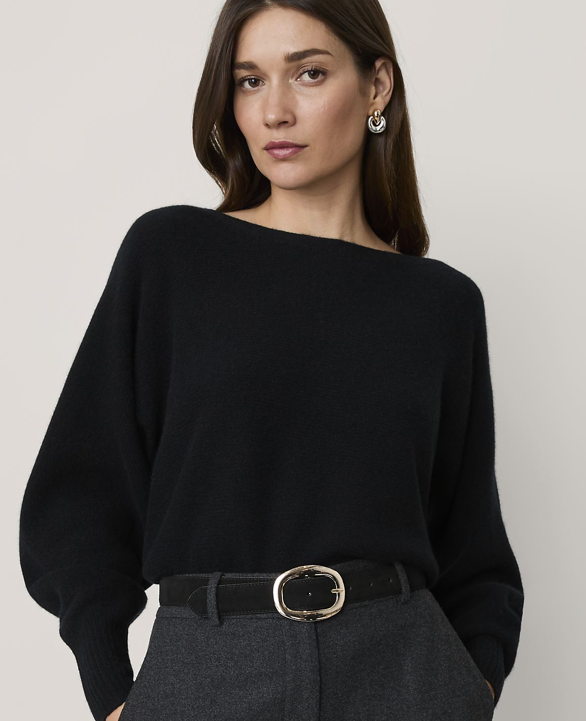 A woman in a black sweater and grey pants stands against a plain background.