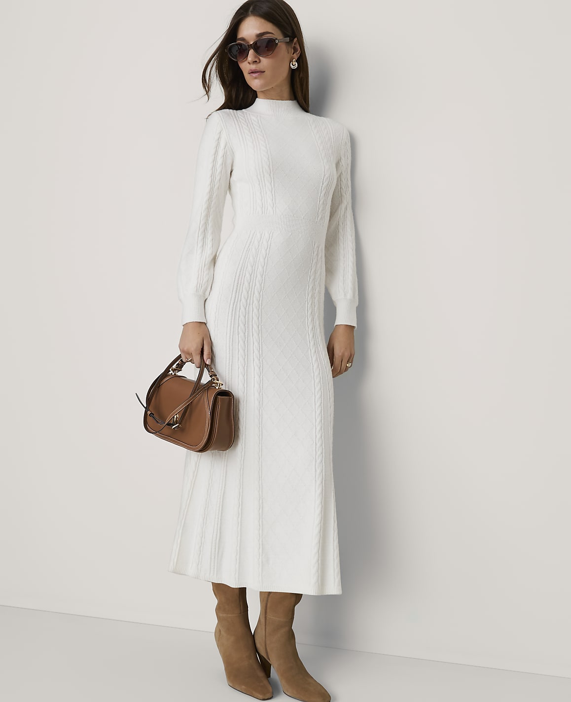 A woman in a white cable-knit dress and brown boots stands against a plain white wall, holding a brown handbag.