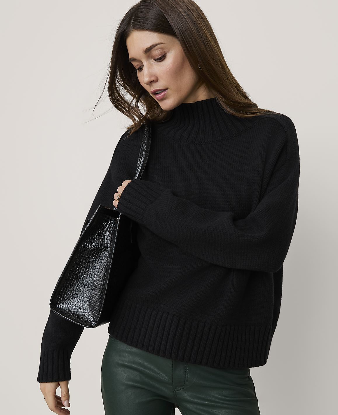 A woman in a black turtleneck sweater and green leather pants carries a black textured handbag against a plain gray background.