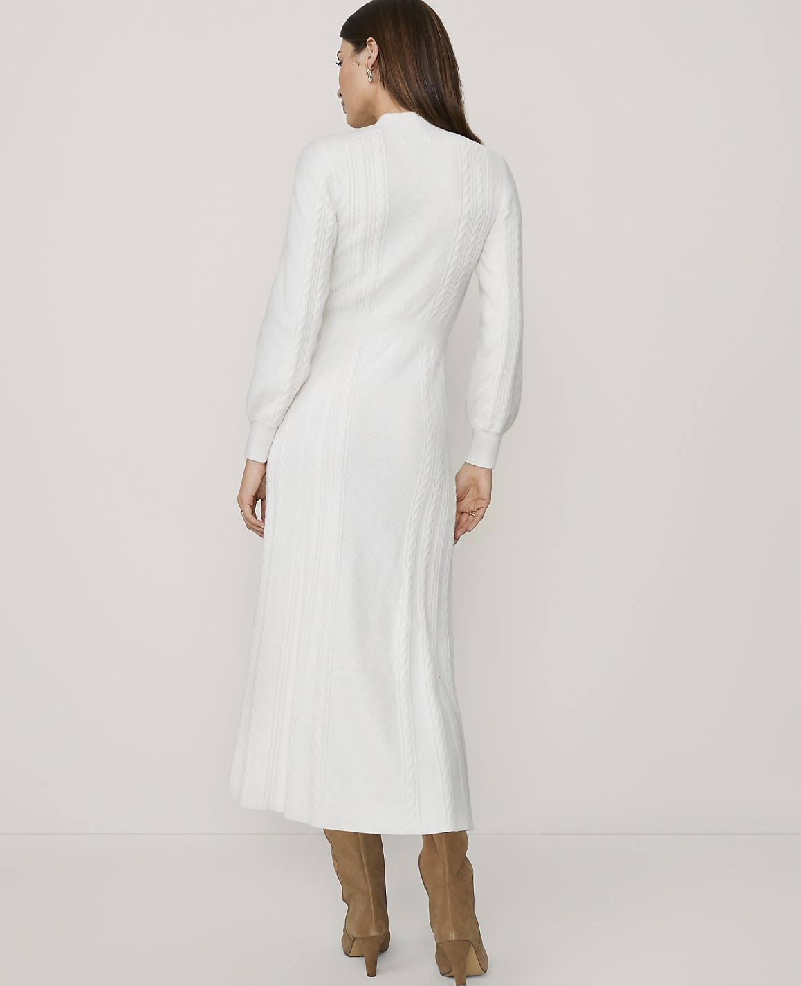 A woman in a white cable-knit dress and brown boots stands against a plain white background.