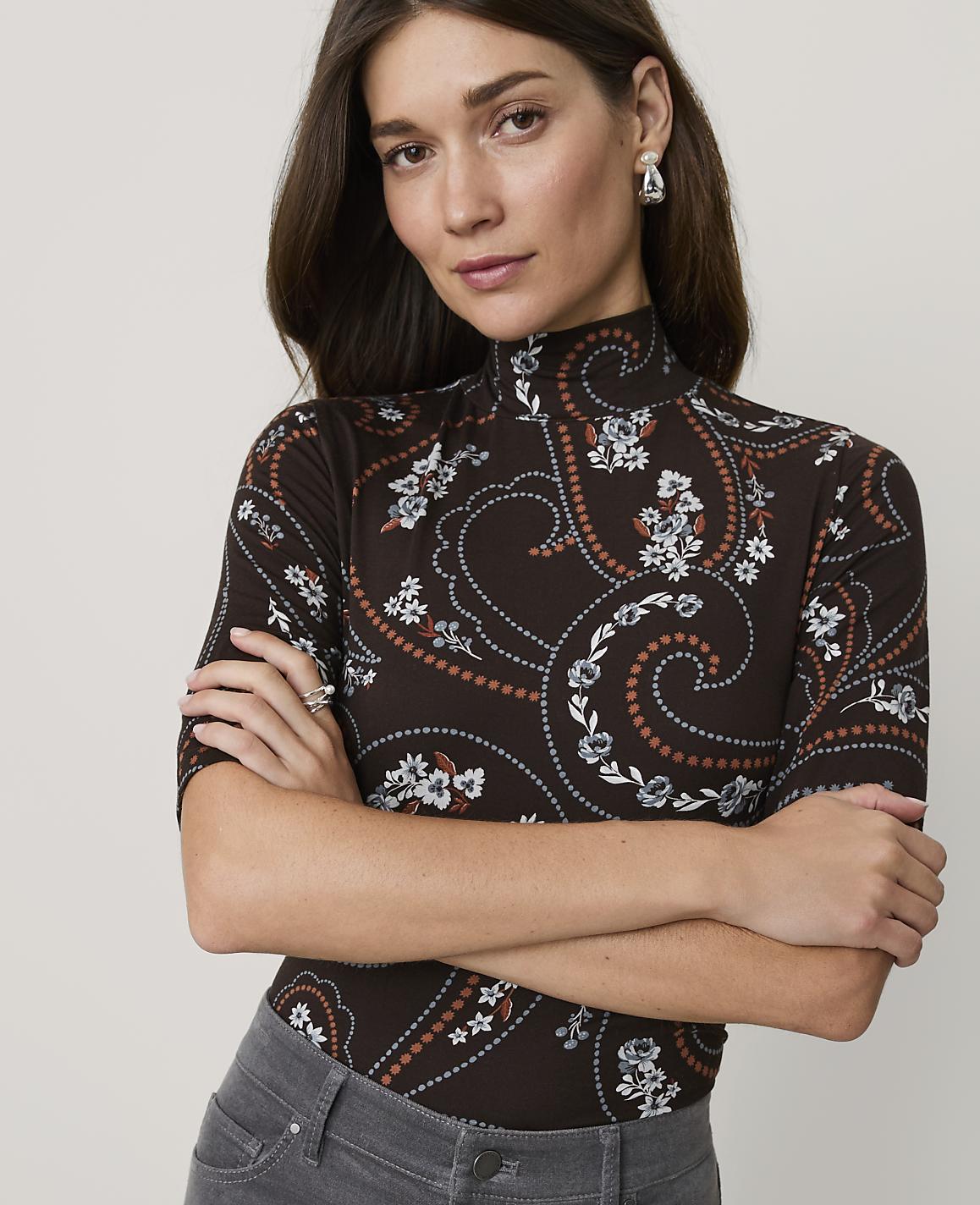 A woman with dark hair and a paisley-patterned turtleneck stands with her arms crossed against a plain background.