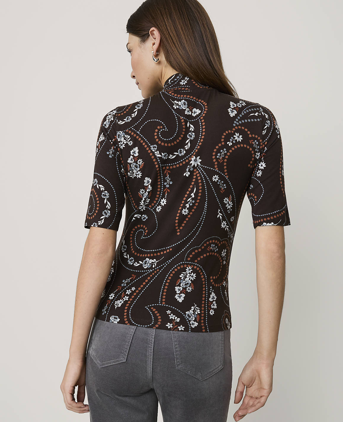 A woman in a brown paisley-patterned top and gray jeans stands against a plain, light-colored background.