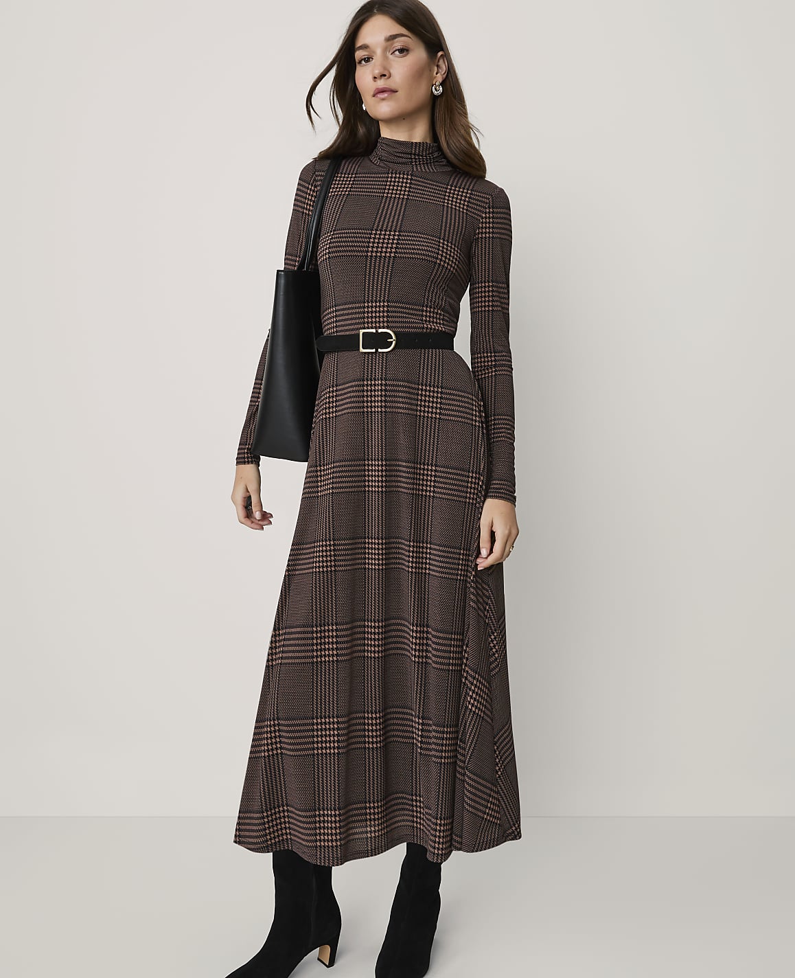 A woman in a brown plaid dress, black belt, and boots stands against a plain background.