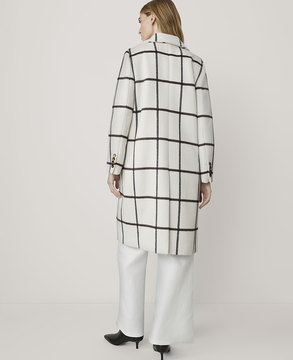 A woman in a white and black checkered coat and white pants stands against a plain white background.