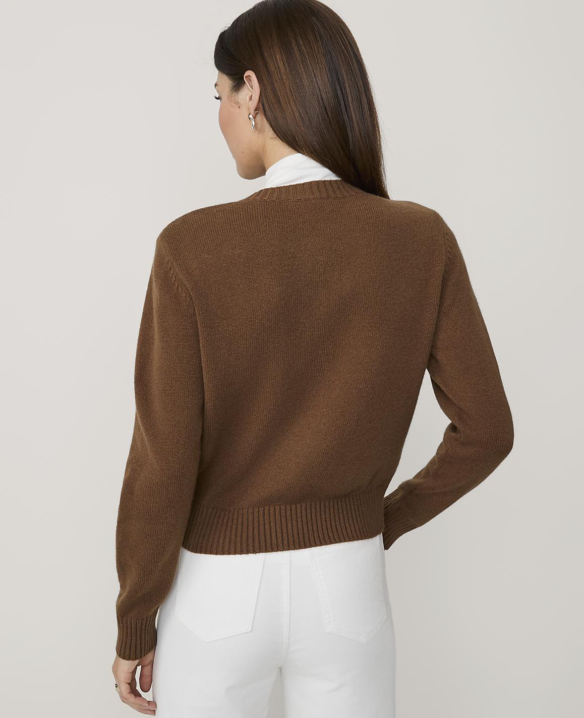 A woman in a brown sweater and white pants stands against a plain white background.