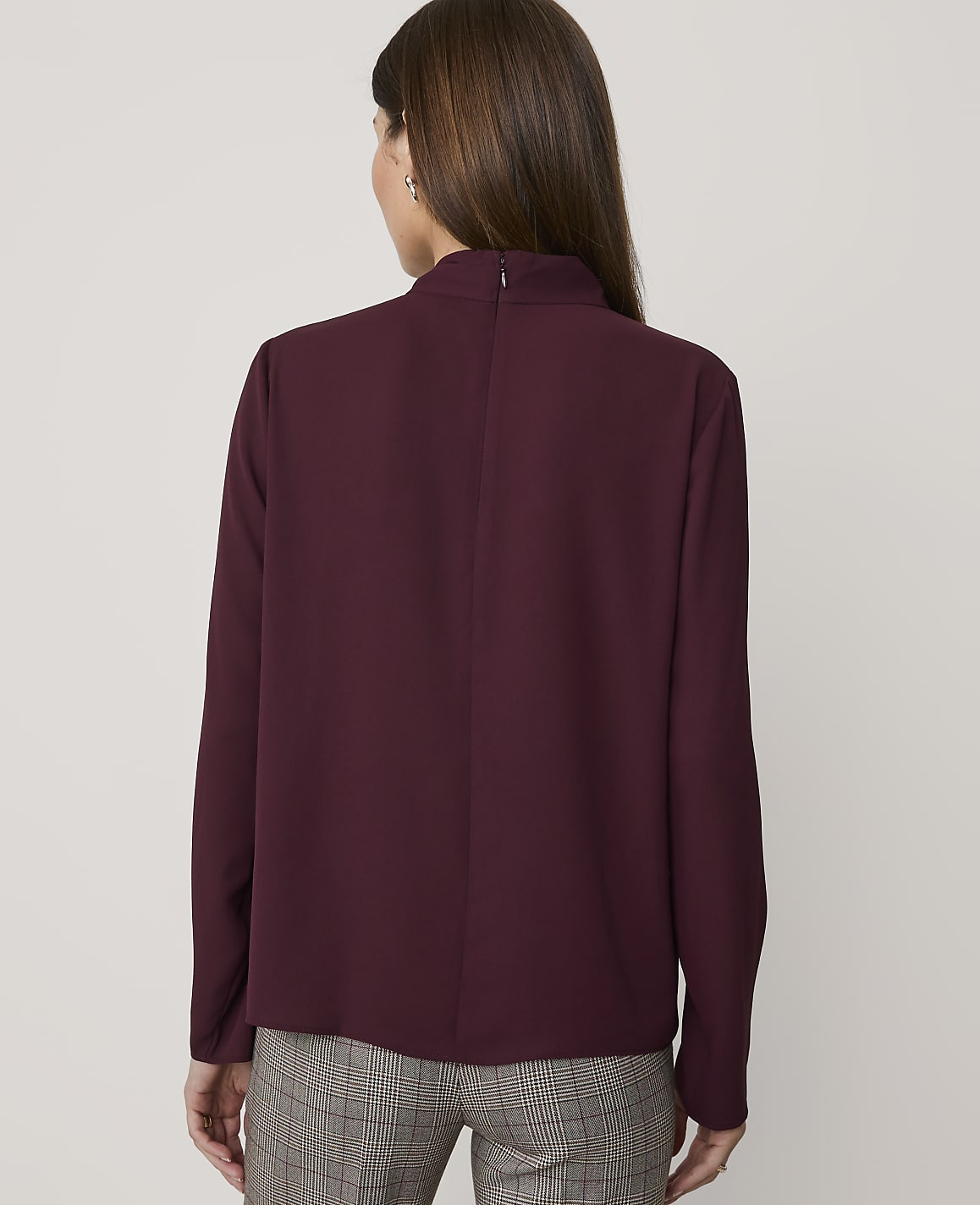 A woman in a burgundy mock-neck blouse and plaid pants stands against a plain gray background.
