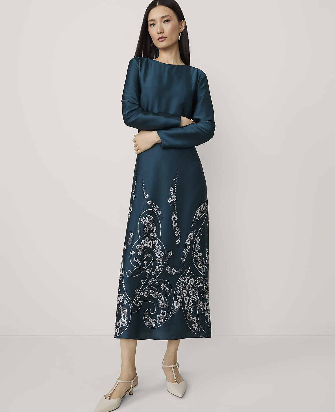 A woman in a teal dress with white floral embroidery stands with her arms crossed against a plain white background.