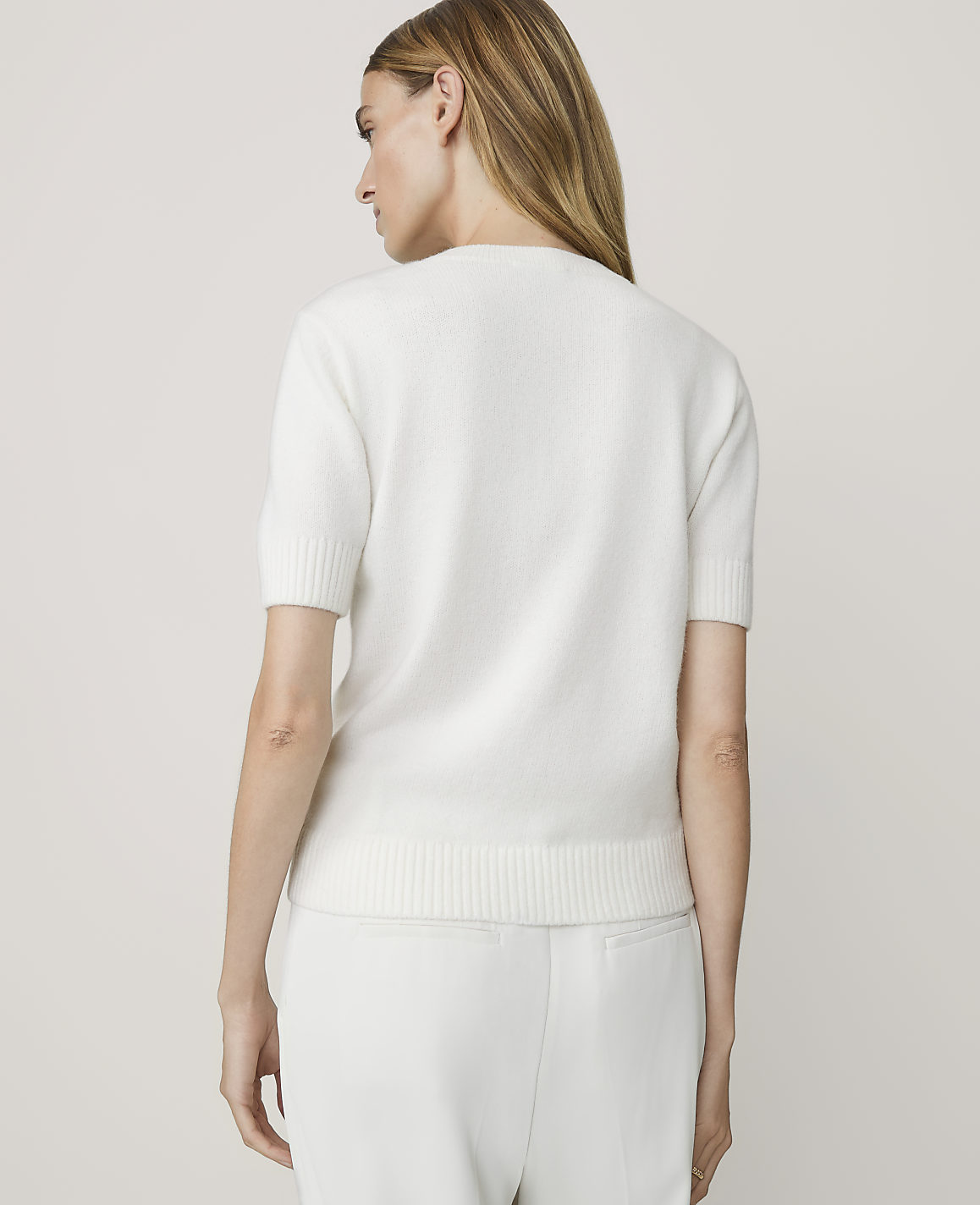 A woman in a white short-sleeved sweater and pants stands against a plain white background.