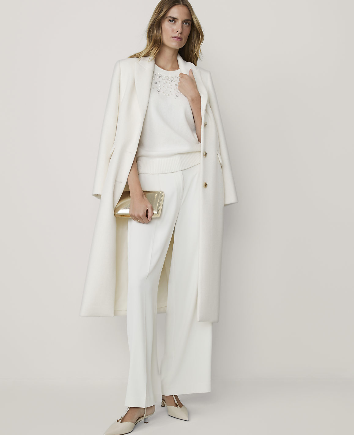 A woman in a white coat and matching pants holds a gold clutch in the foreground, against a plain white background.