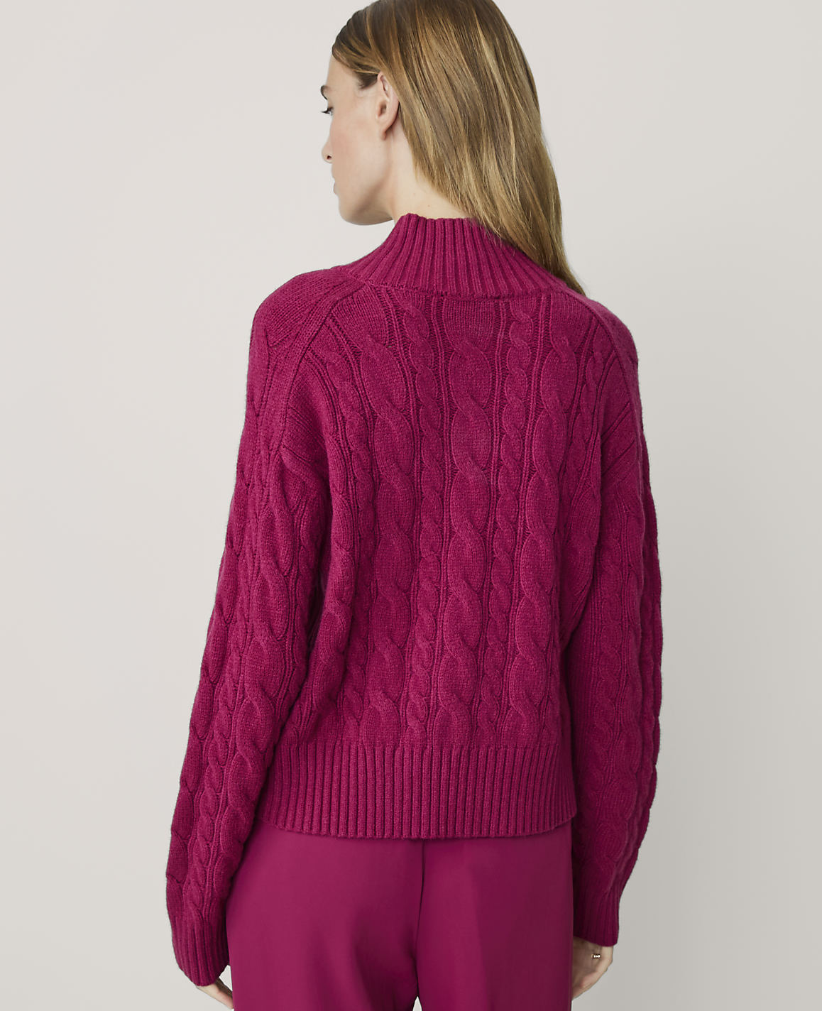 A woman in a magenta cable-knit sweater and matching pants stands against a plain, light background.