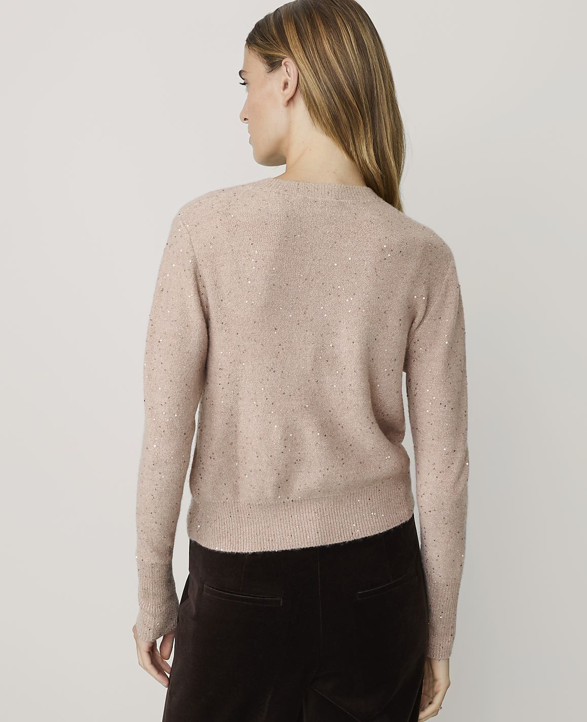 A woman in a sparkly pink cardigan and dark brown pants stands against a plain, light-colored background.