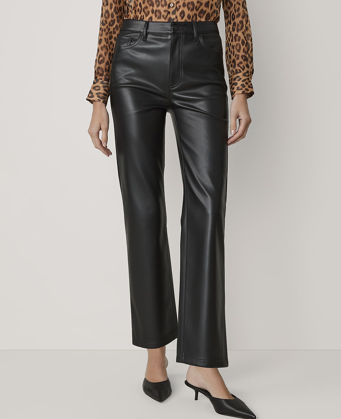 A person wearing black faux leather pants and a leopard print blouse stands against a plain white background.