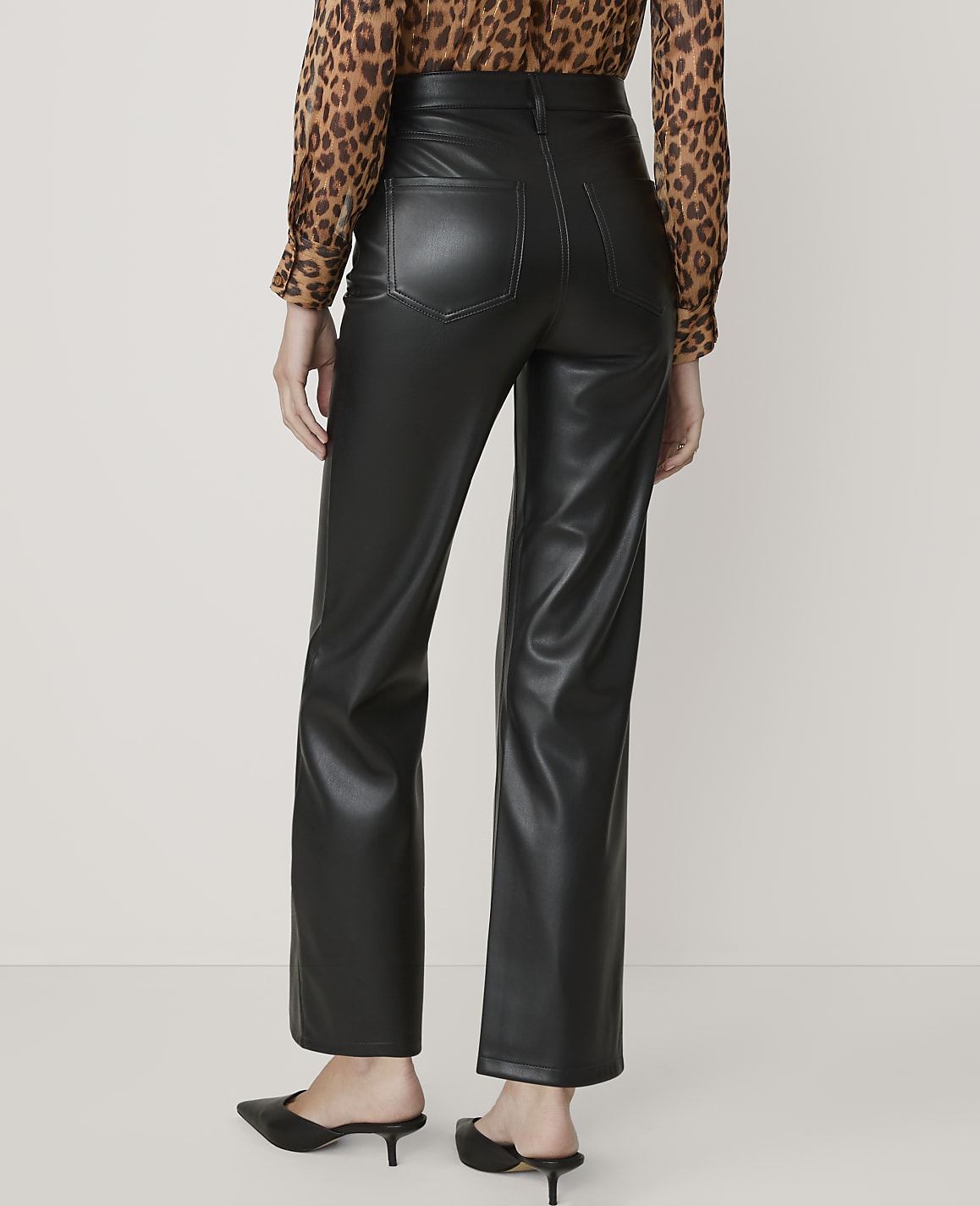 A person wearing black faux leather pants and a leopard print blouse stands against a plain, light-colored background.