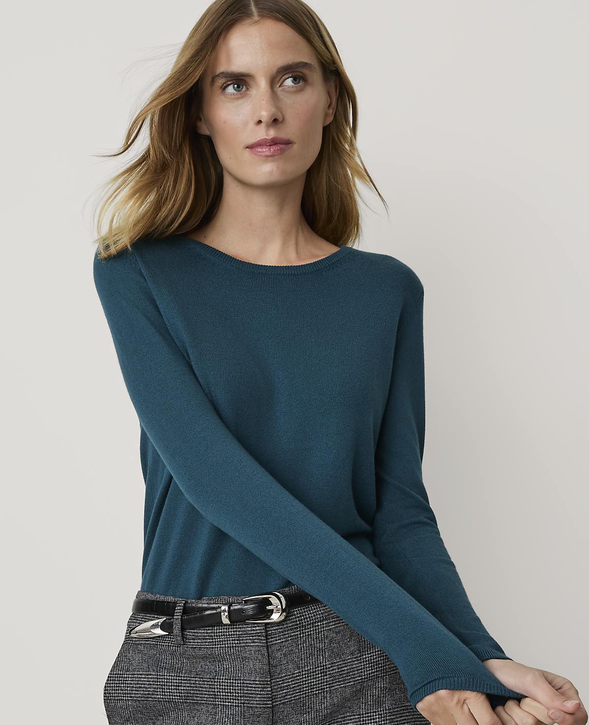 A woman in a teal sweater and grey pants stands against a plain white background.