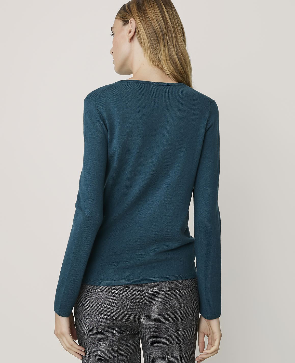A woman in a teal sweater and patterned pants stands against a plain, light-colored background.