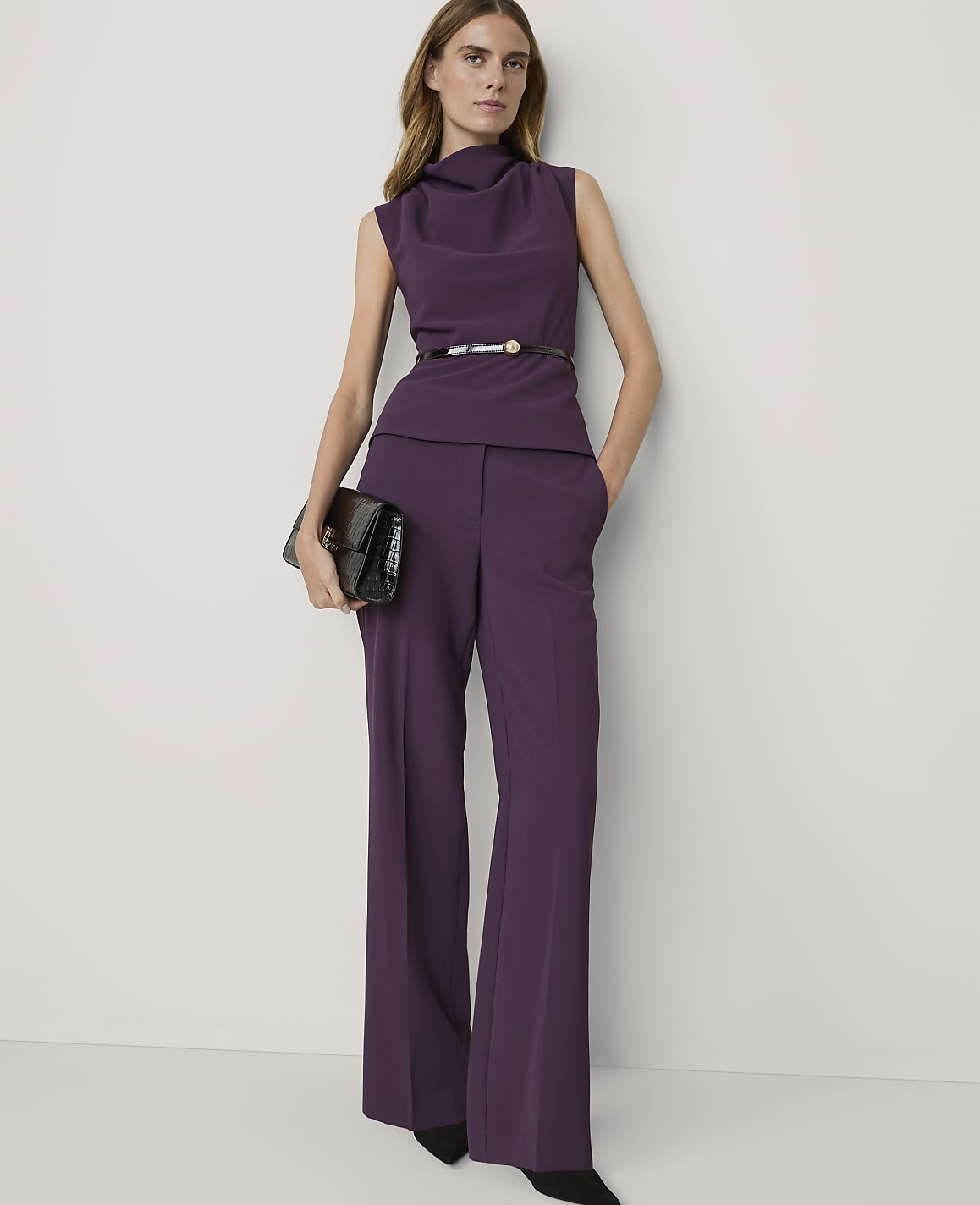 A woman in a purple pantsuit with a cowl neck and a black clutch stands against a plain white background.