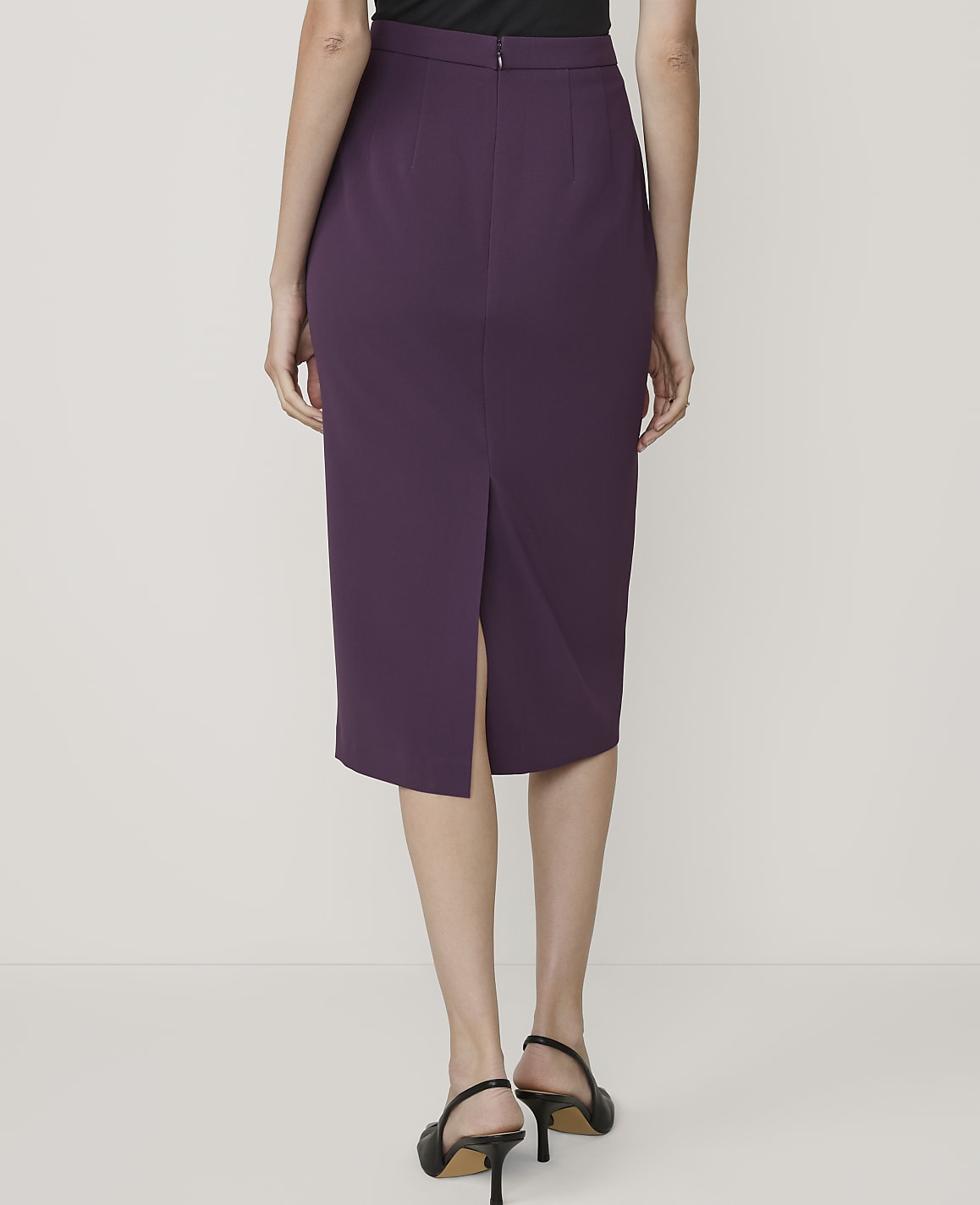 A woman in a purple pencil skirt and black heels stands against a plain white background.