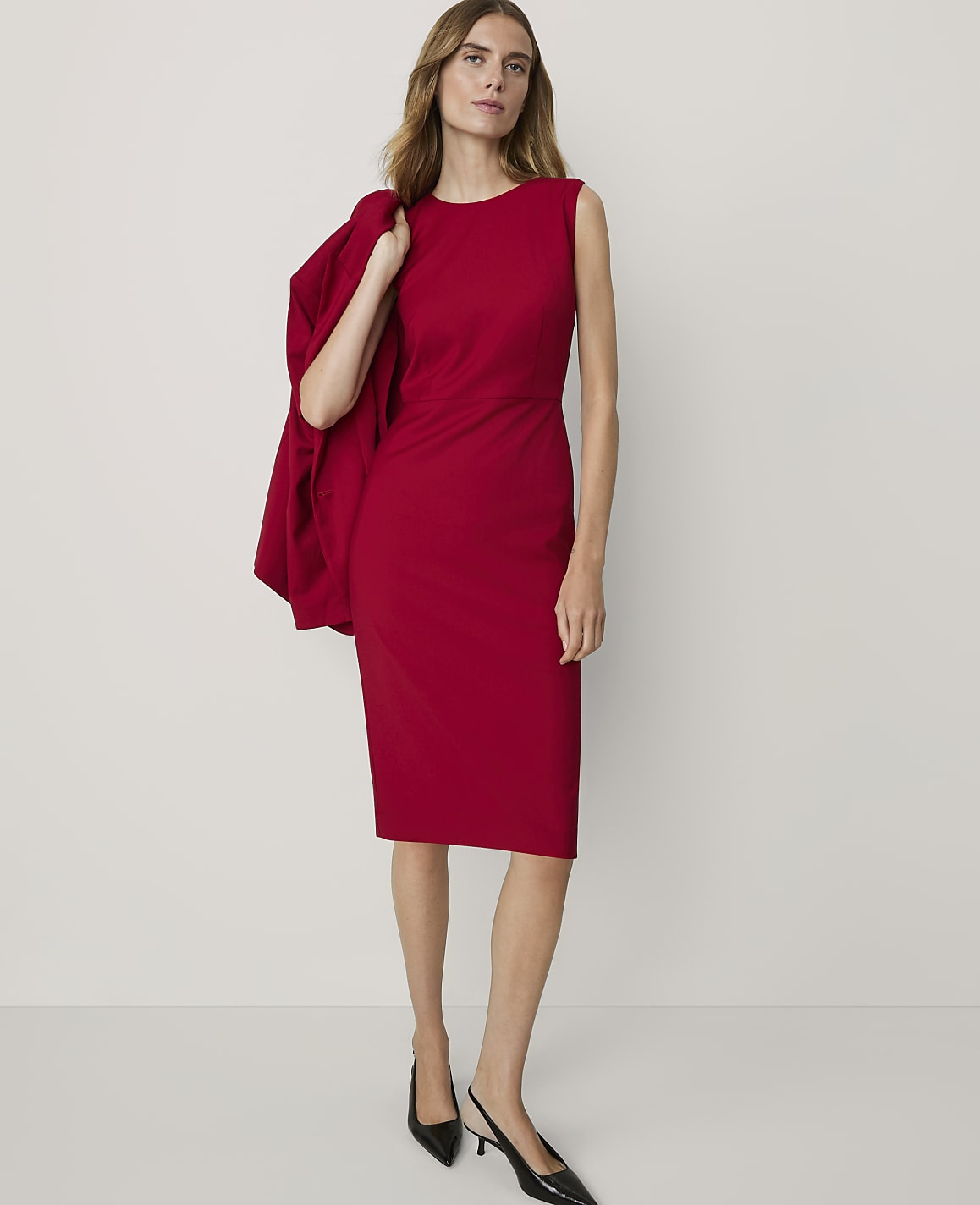 A woman in a red dress and black heels stands against a plain white background, with a red blazer draped over her shoulder.