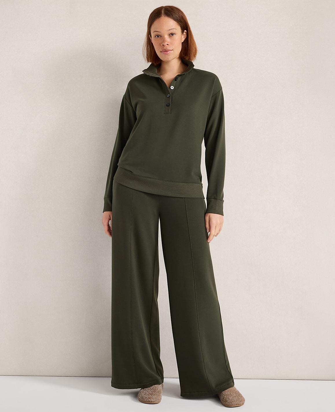 A woman stands in a dark green loungewear set with wide-leg pants and a collared top, against a plain, textured wall.