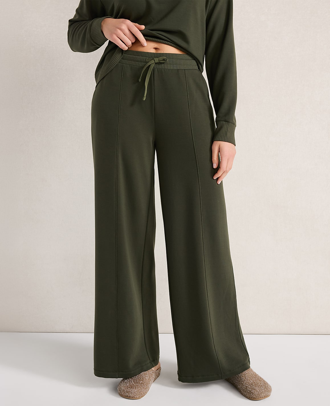 A person in olive green wide-leg pants and slippers stands against a neutral, textured background.