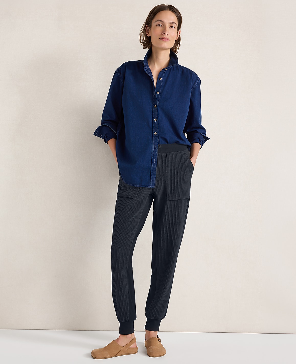 A woman in a dark blue shirt and striped pants stands against a textured cream wall.