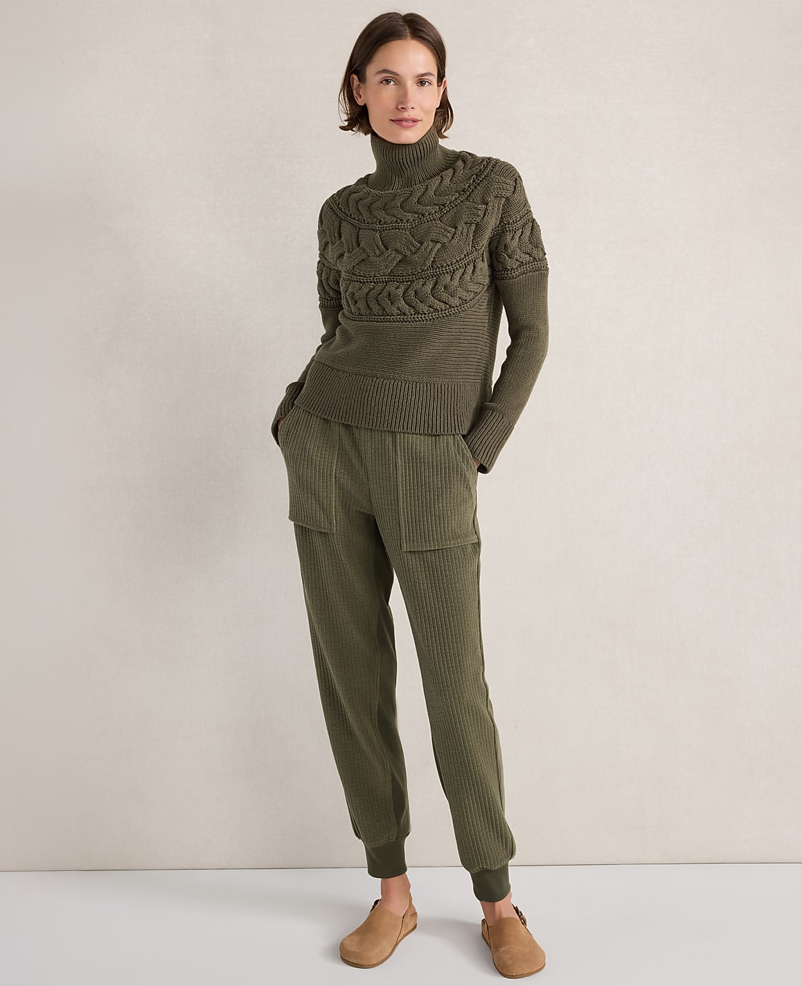 A woman in a green cable-knit turtleneck sweater and matching ribbed pants stands against a plain, light brown background.